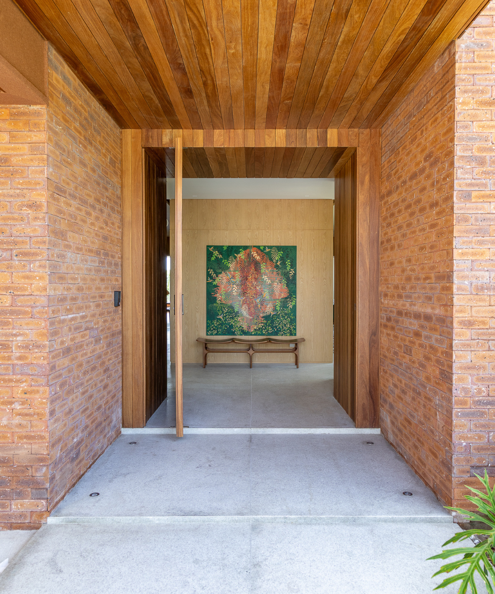 Entrada com parede de tijolinho aparente em tons terrosos, porta pivotante e teto em madeira, criando ambiente acolhedor e
