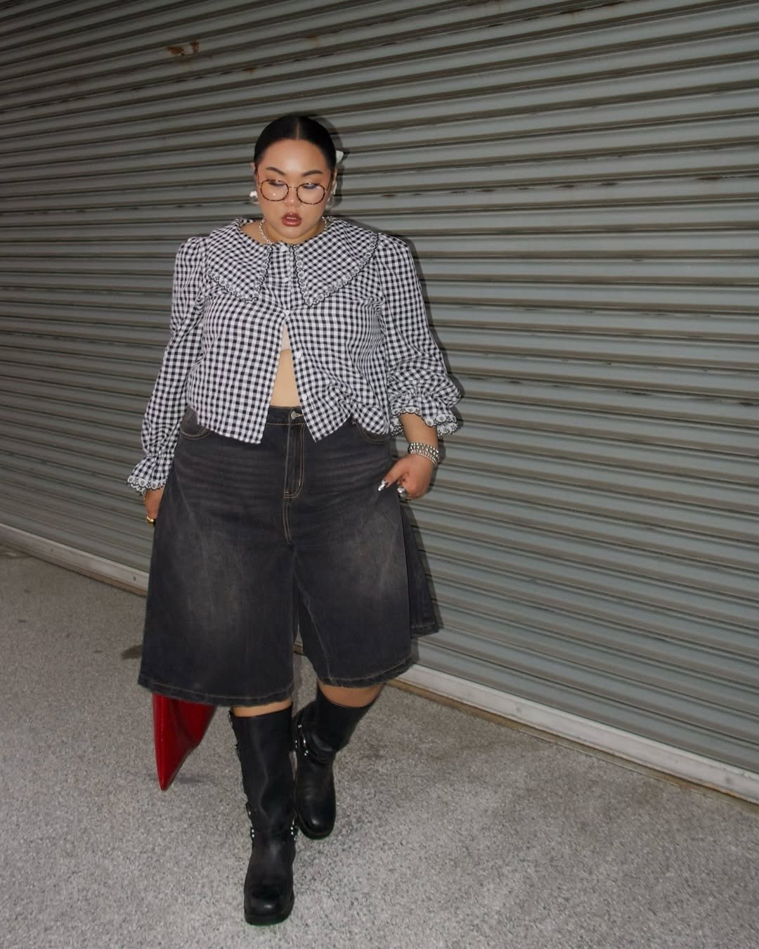 Modelo caminha com jorts preto de jeans largo, blusa xadrez preta e branca com gola oversized e botas pretas.