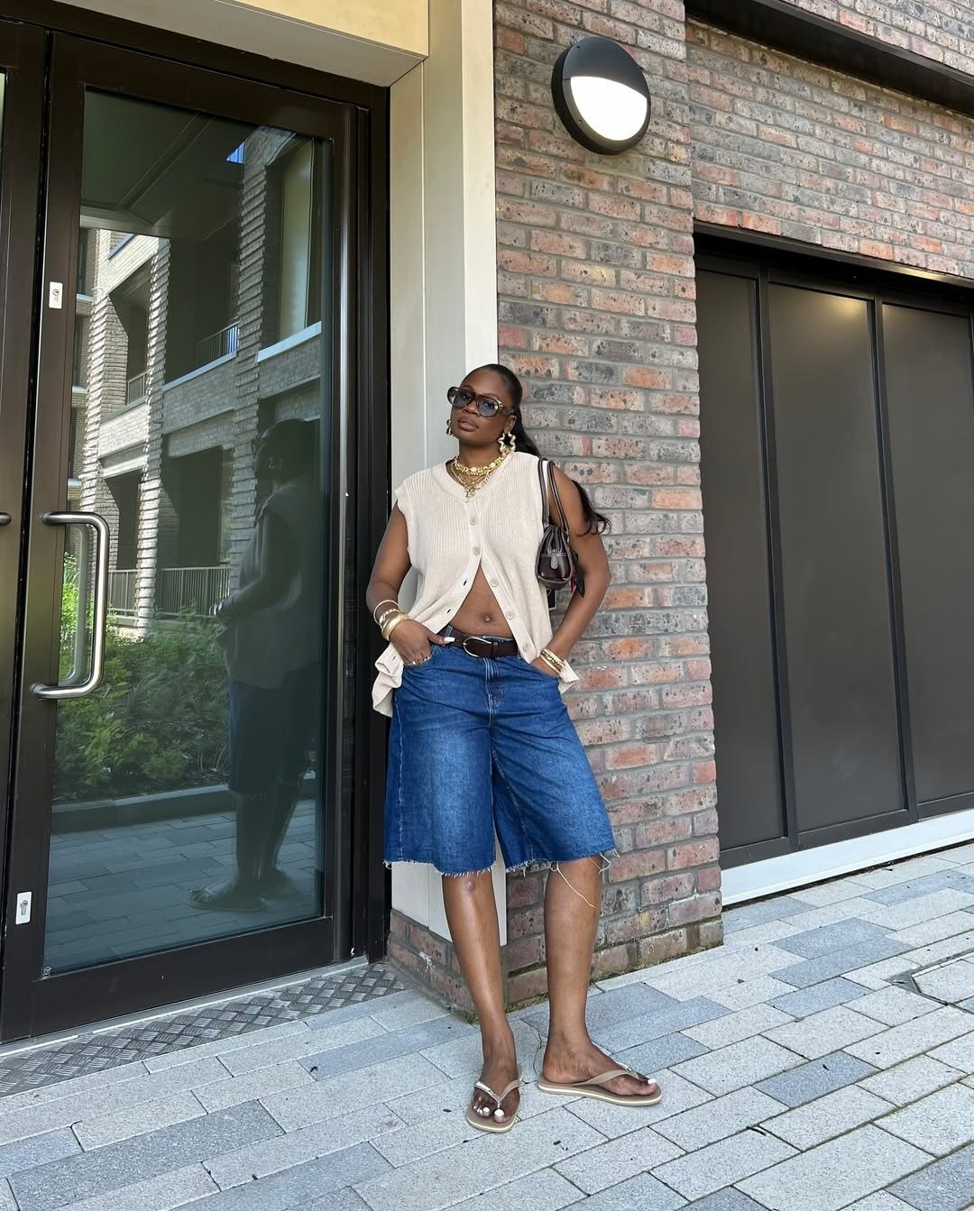 Modelo posa com jorts jeans azul de comprimento médio e barra desfiada, combinando colete bege de tricô e acessórios