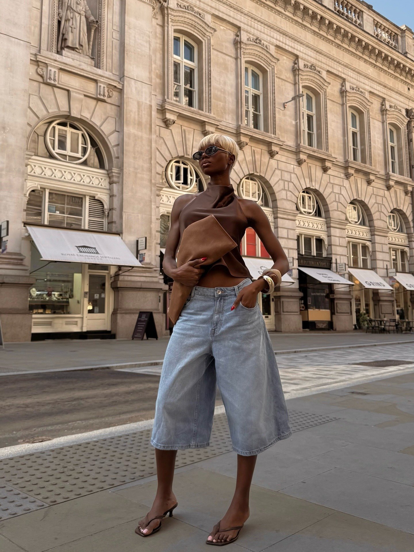 Modelo posa confiante com jorts largos de jeans claro, blusa marrom de tecido acetinado e acessórios dourados.