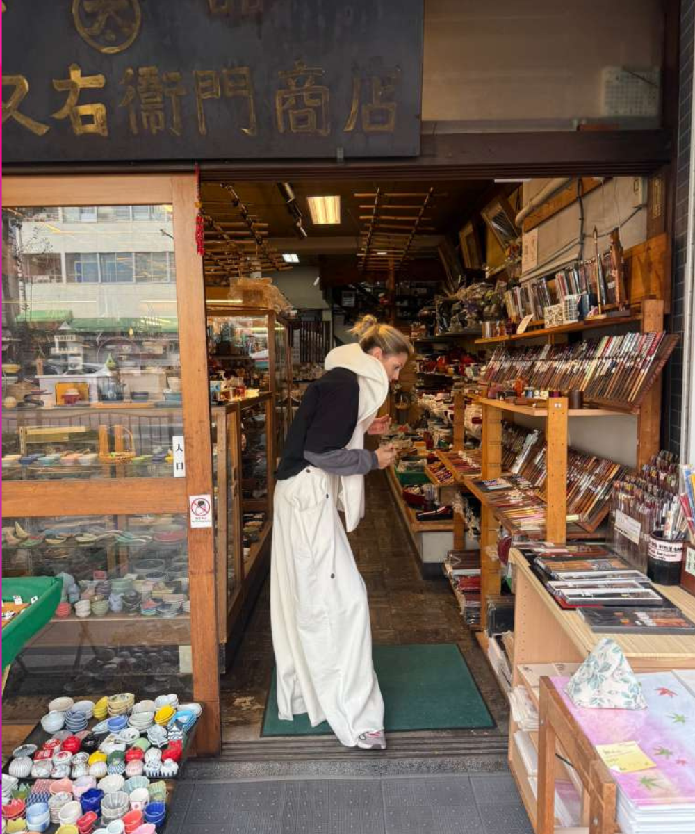 Mulher com calça branca ampla, blusa preta e cachecol branco explora loja tradicional em Tóquio, destacando dicas de Tóquio
