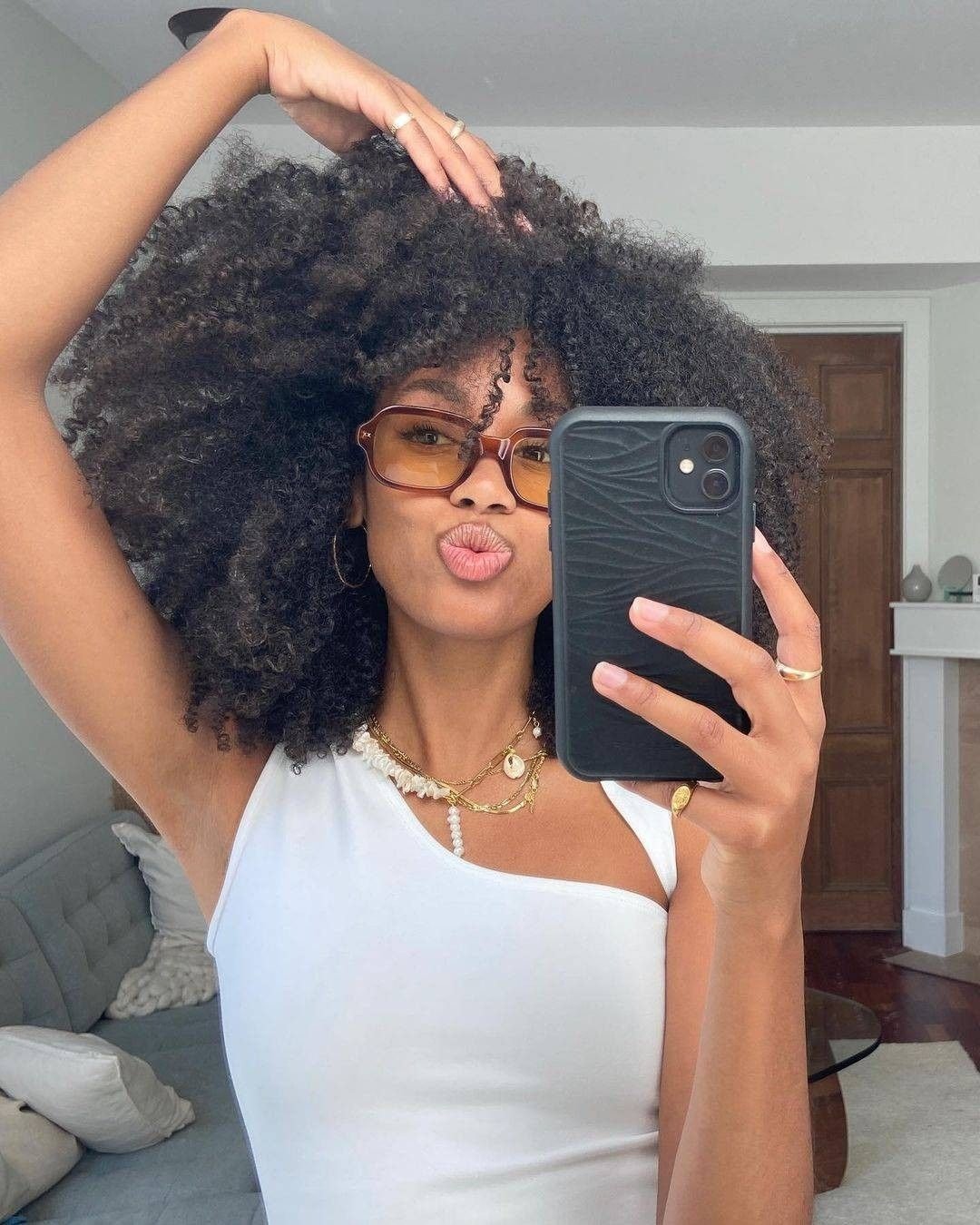 Modelo posa para selfie com corte de cabelo 2026 volumoso e cacheado, usando blusa branca assimétrica e acessórios dourados