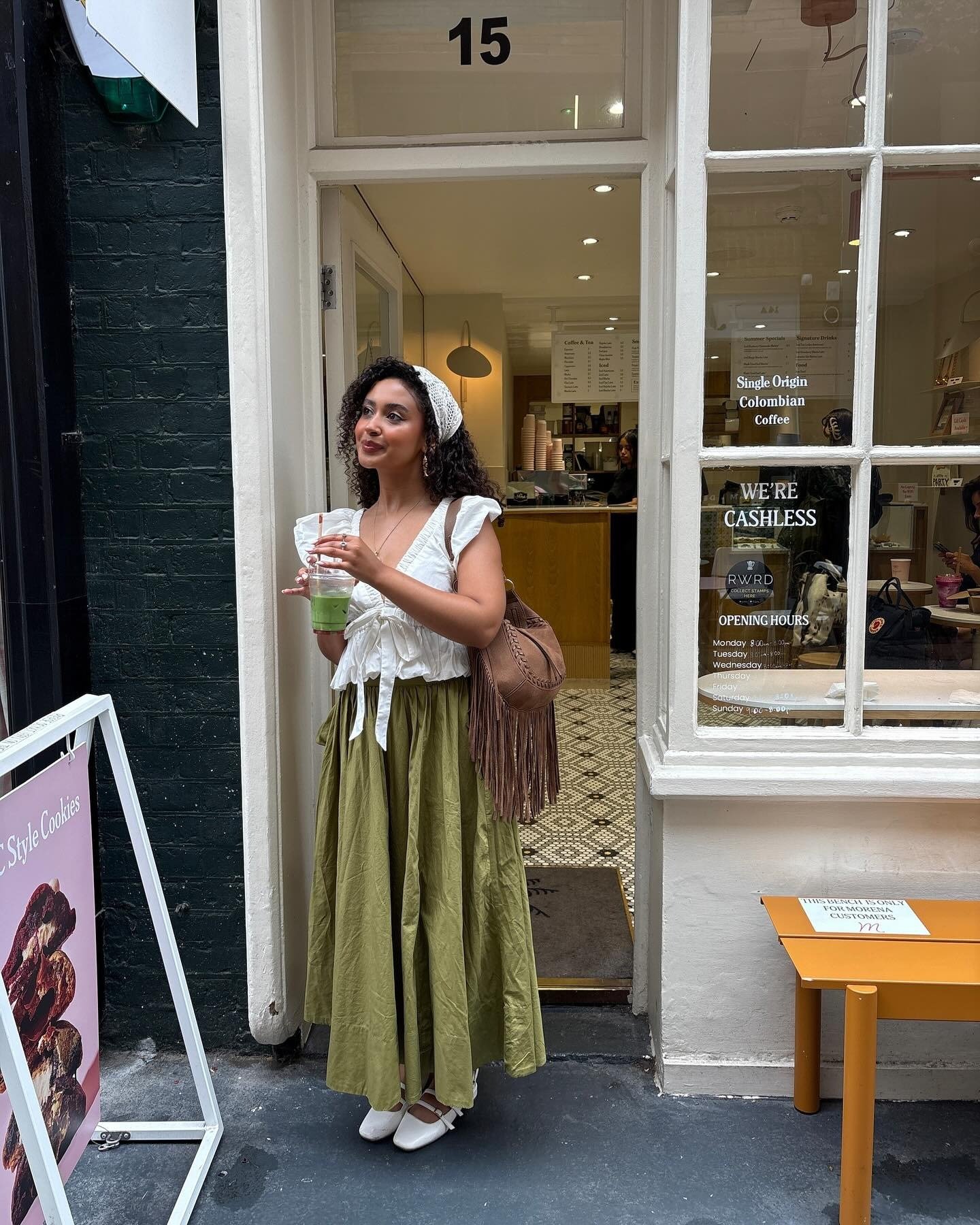 Modelo posa com blusa branca de mangas bufantes e amarração frontal, saia longa verde em tecido leve e bolsa marrom