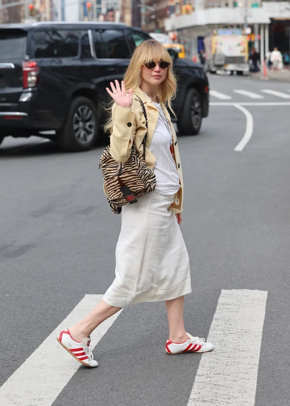 Modelo caminha na rua com blazer bege, camiseta branca, saia midi de linho e tênis branco com listras vermelhas.