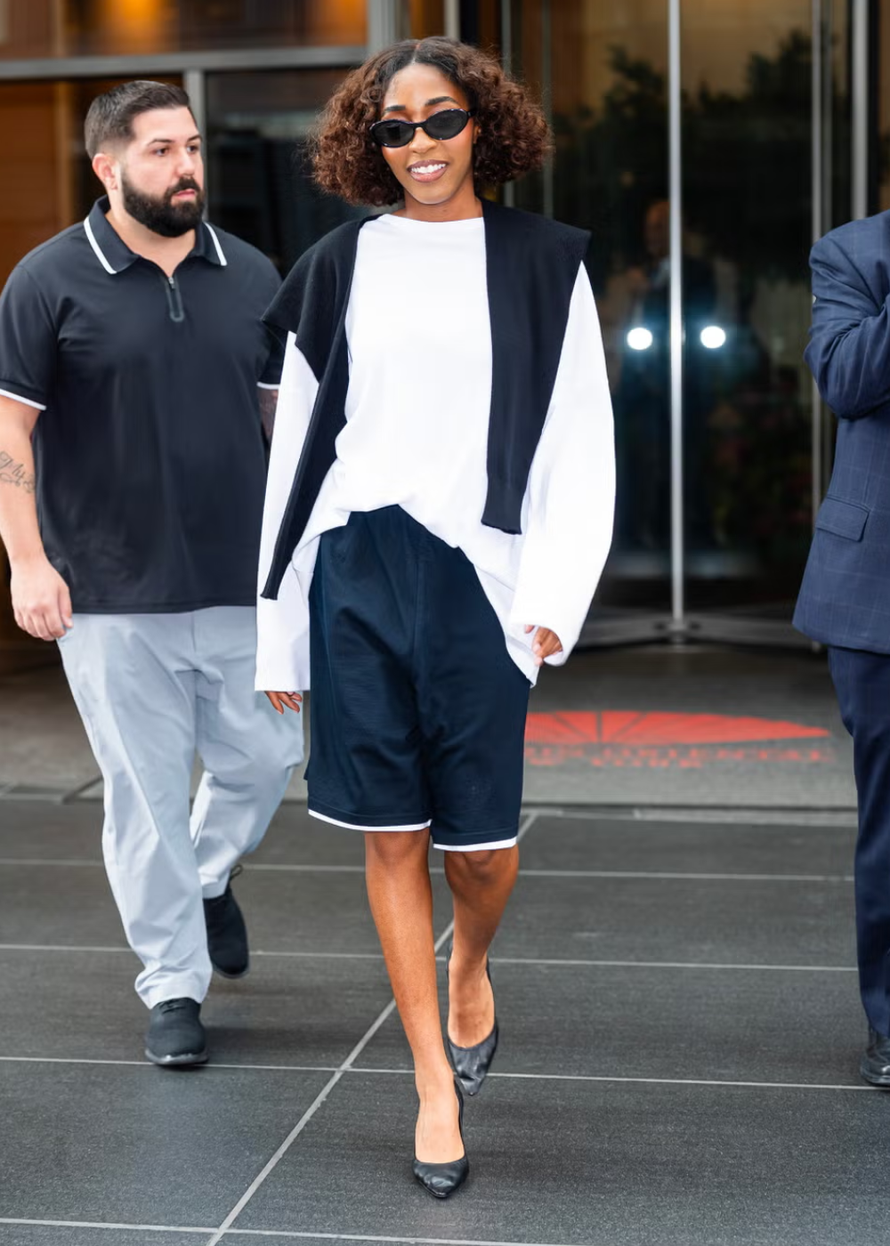 Modelo caminha confiante com looks básicos em camiseta branca oversized, bermuda preta de malha e scarpin preto, com suéter