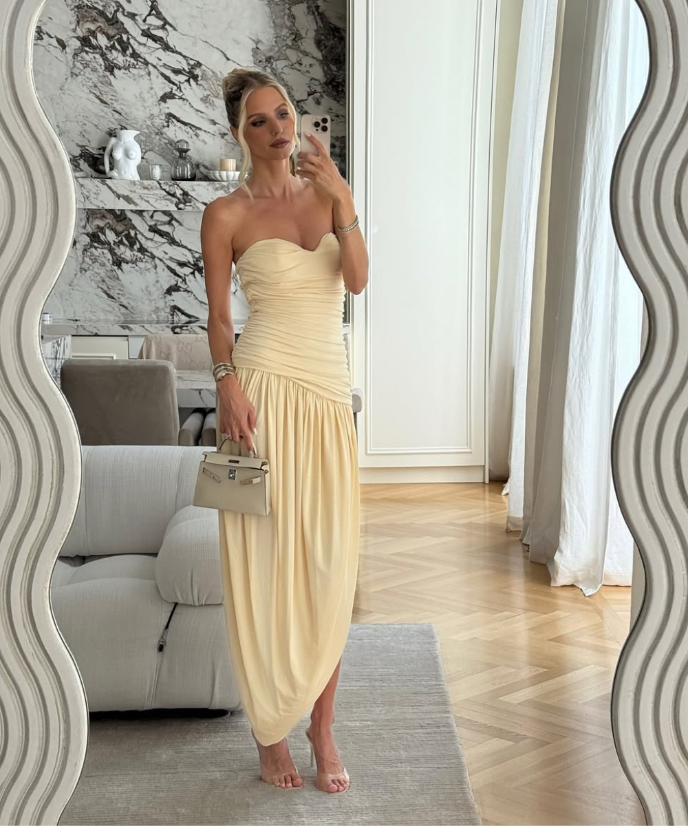 Modelo posa em vestido tomara que caia amarelo claro com drapeados e saia fluida, combinando bolsa nude.