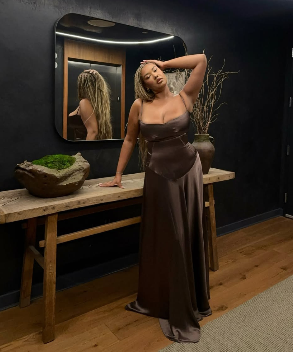 Modelo posa com vestido longo de cetim marrom, decote quadrado e caimento fluido, em ambiente sofisticado e acolhedor,