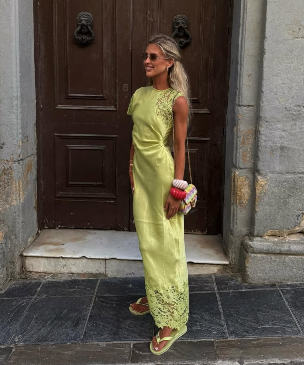 Modelo posa sorrindo com vestido longo verde claro de tecido leve e detalhes em renda, combinando sandálias e acessórios.