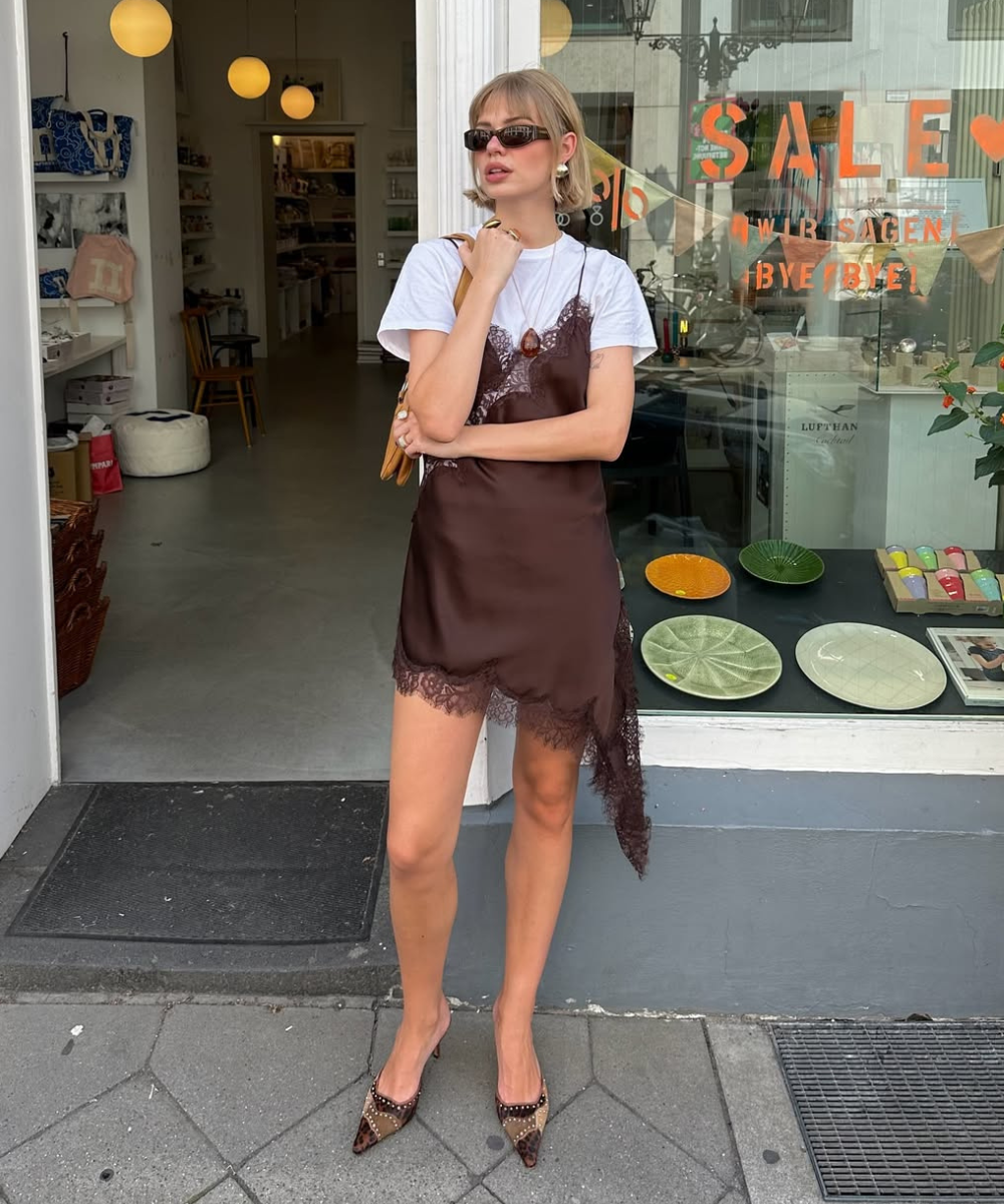 Modelo posa com vestido slip dress marrom de cetim e renda sobre camiseta branca oversized, combinando scarpin animal print.