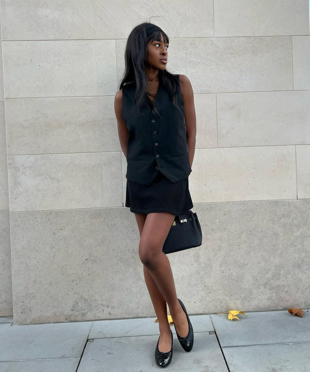 Modelo posa com look estiloso em preto total, combinando colete estruturado, saia curta e sapatilhas de verniz.