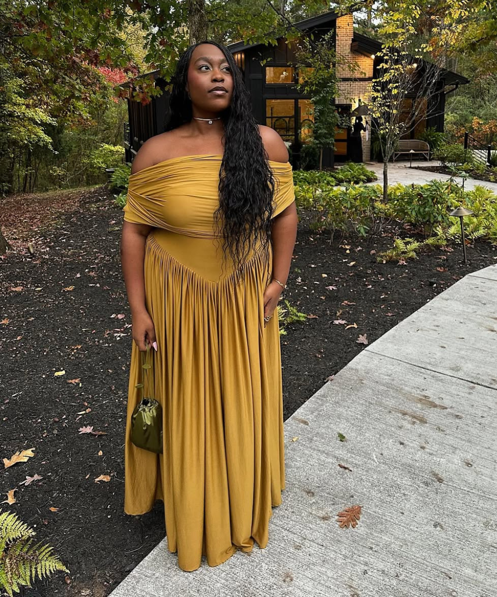 Modelo posa com vestido longo mostarda de tecido fluido e ombros à mostra, combinando bolsa de cetim verde.