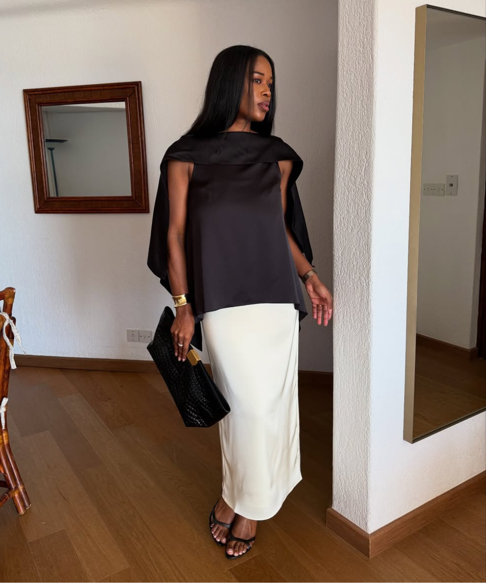 Modelo posa com blusa preta de tecido acetinado e saia longa branca, combinando clutch texturizada e sandálias finas.