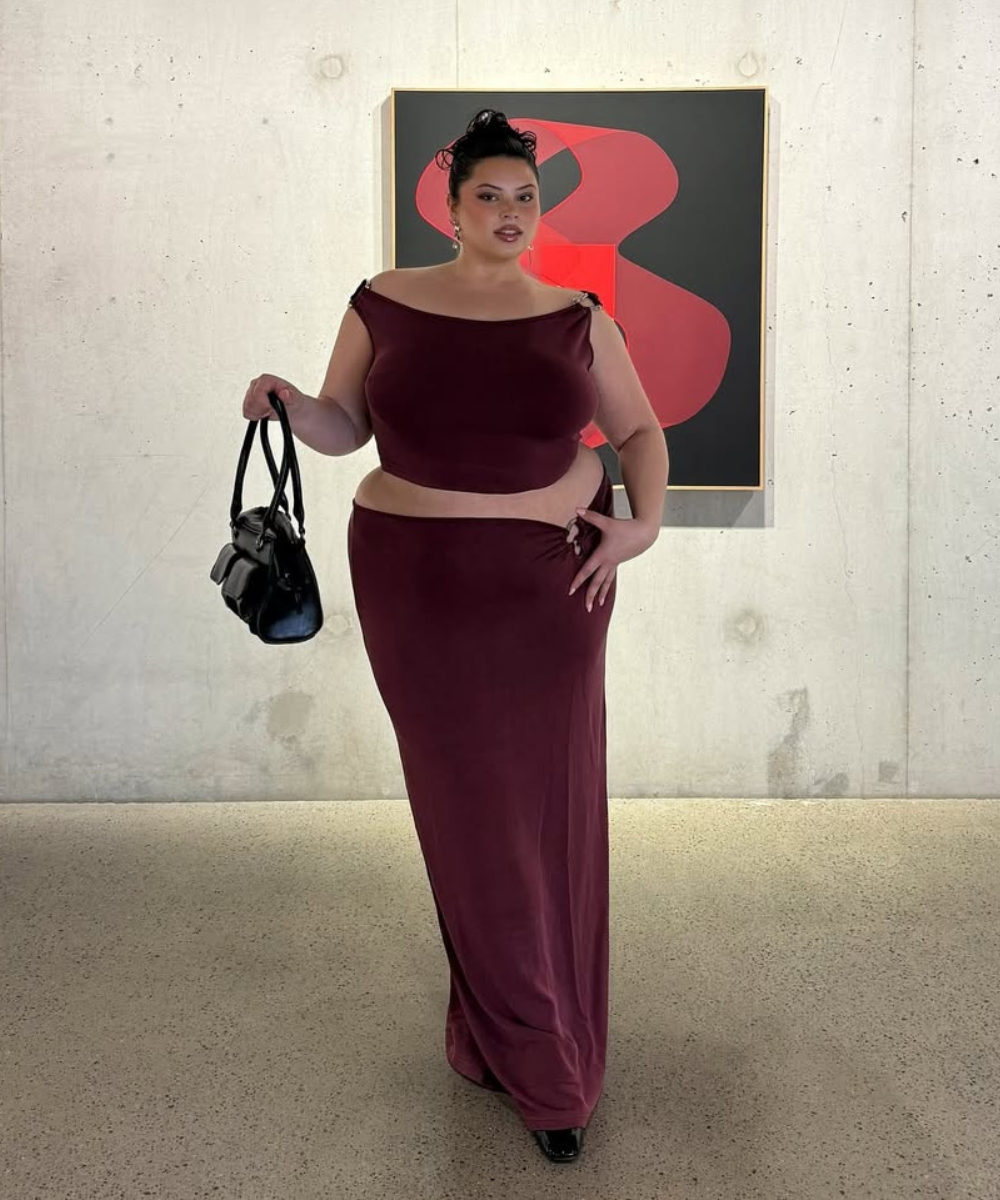 Modelo posa confiante com conjunto vinho de cropped ombro a ombro e saia longa de tecido fluido, combinando bolsa preta.