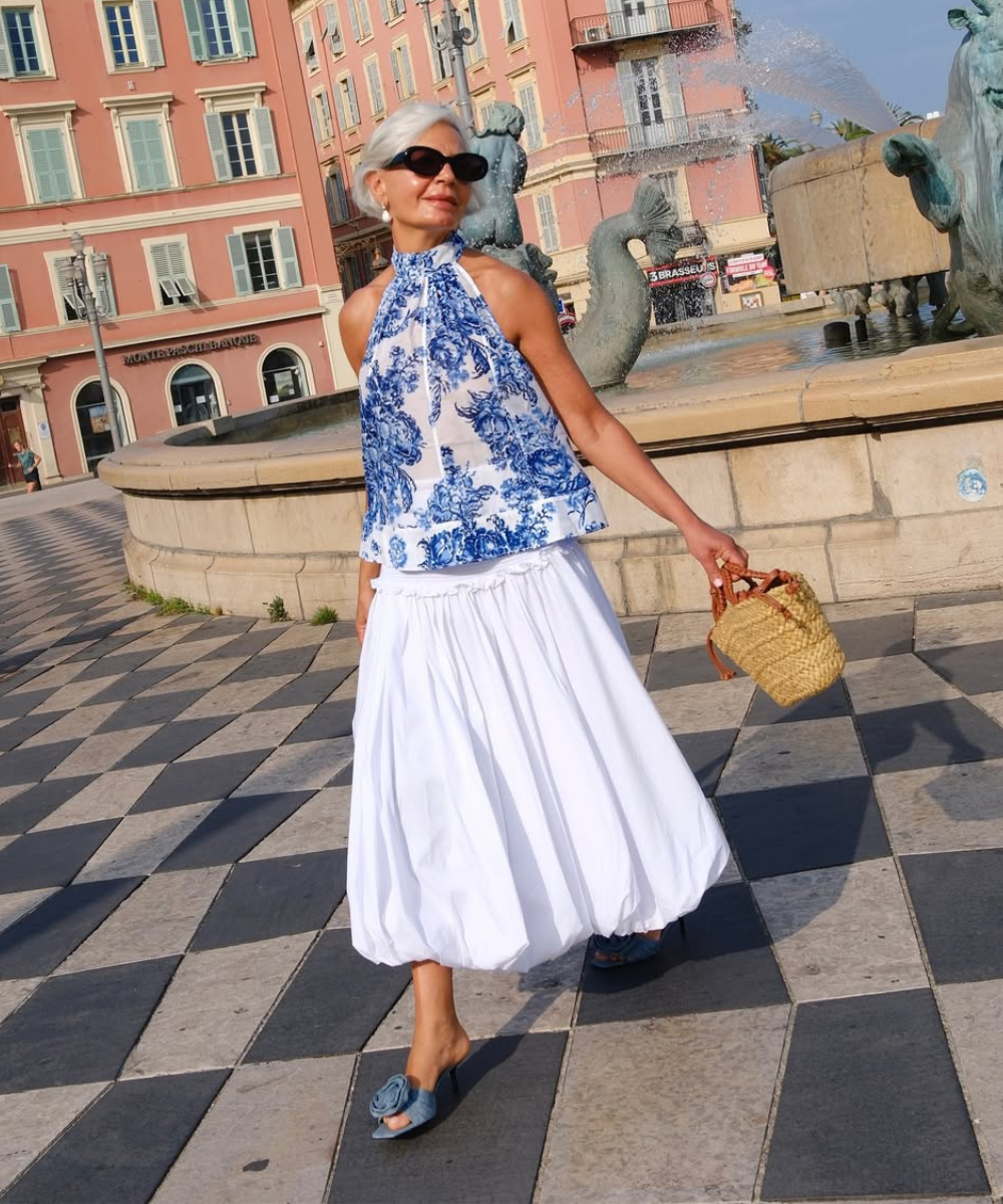Mulher elegante em looks com azul, blusa halter estampada, saia branca ampla e sandálias jeans, em pose confiante e leve.
