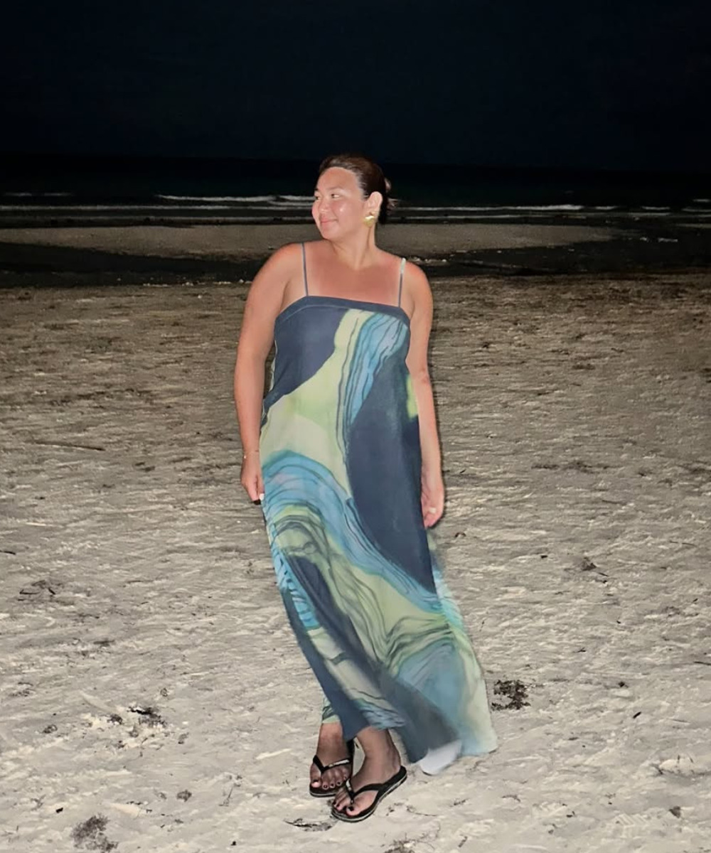 Mulher posa na praia à noite com vestido longo fluido em tons de azul e verde, combinando leveza e elegância.