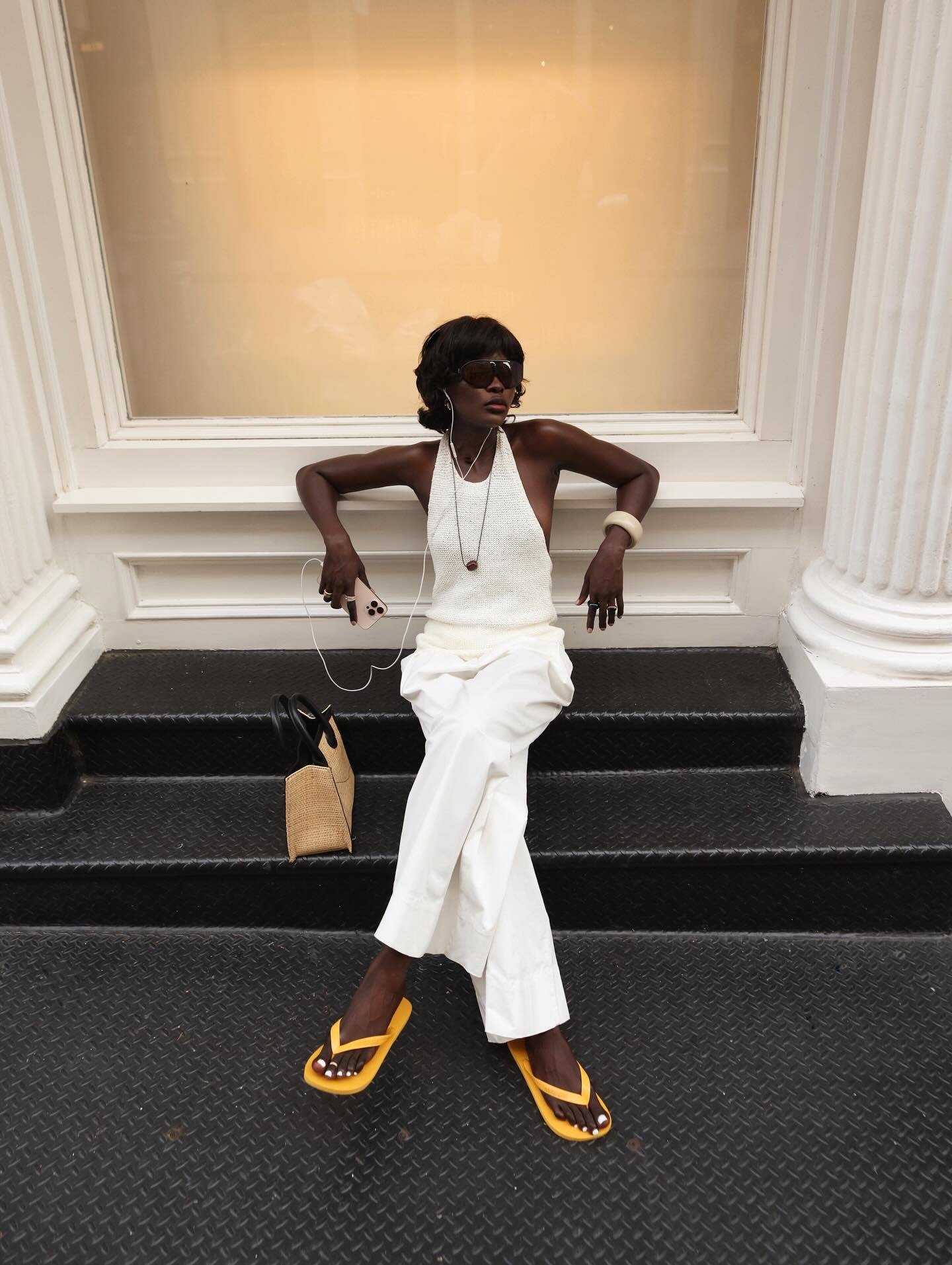 Modelo senta com postura relaxada e segura, usando chinelo amarelo vibrante em borracha, calça branca ampla e regata