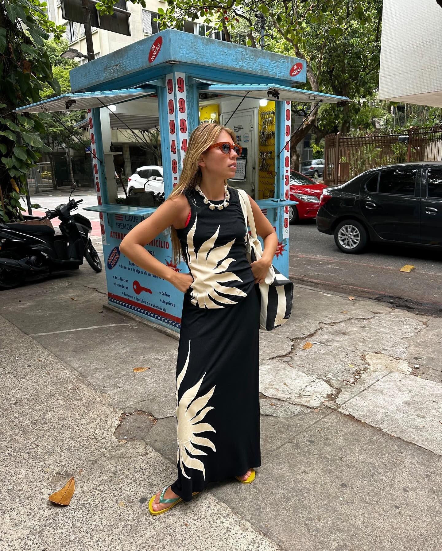 Mulher posa confiante com vestido longo preto estampado em bege e chinelo amarelo de tiras verdes, em cenário urbano.