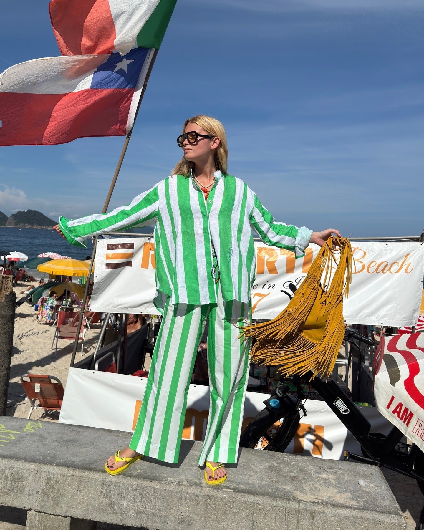 Modelo posa de braços abertos com conjunto listrado verde e branco, chinelo amarelo vibrante e bolsa de franjas mostarda à