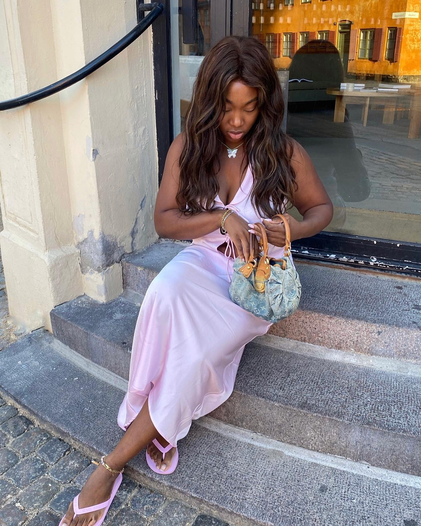 Mulher sentada em escada usa vestido rosa de tecido fluido e cor de chinelo rosa.