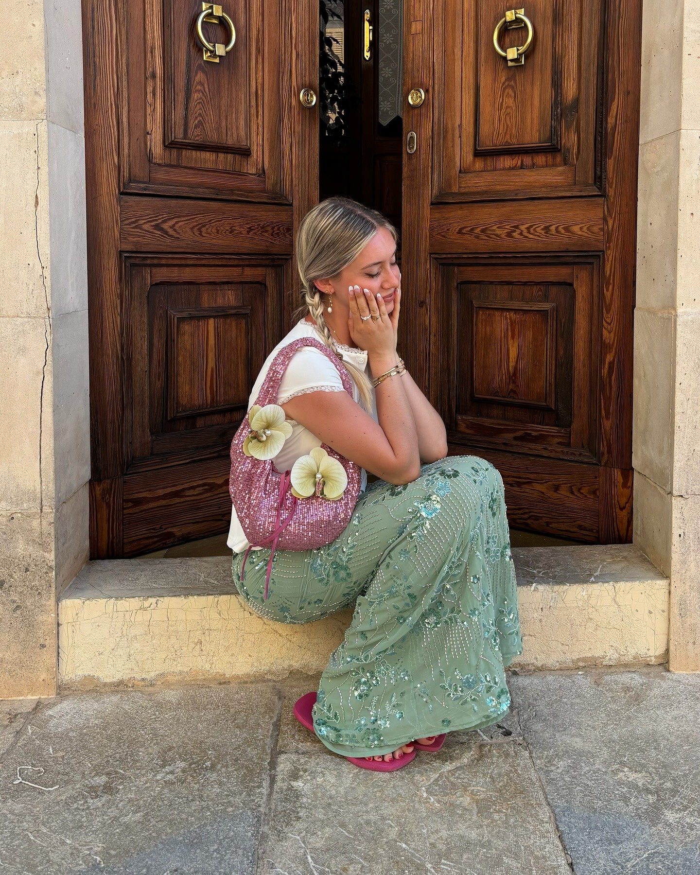 Mulher sentada com saia verde bordada fluida, camiseta branca de manga curta, bolsa rosa com paetês e chinelo rosa casual,