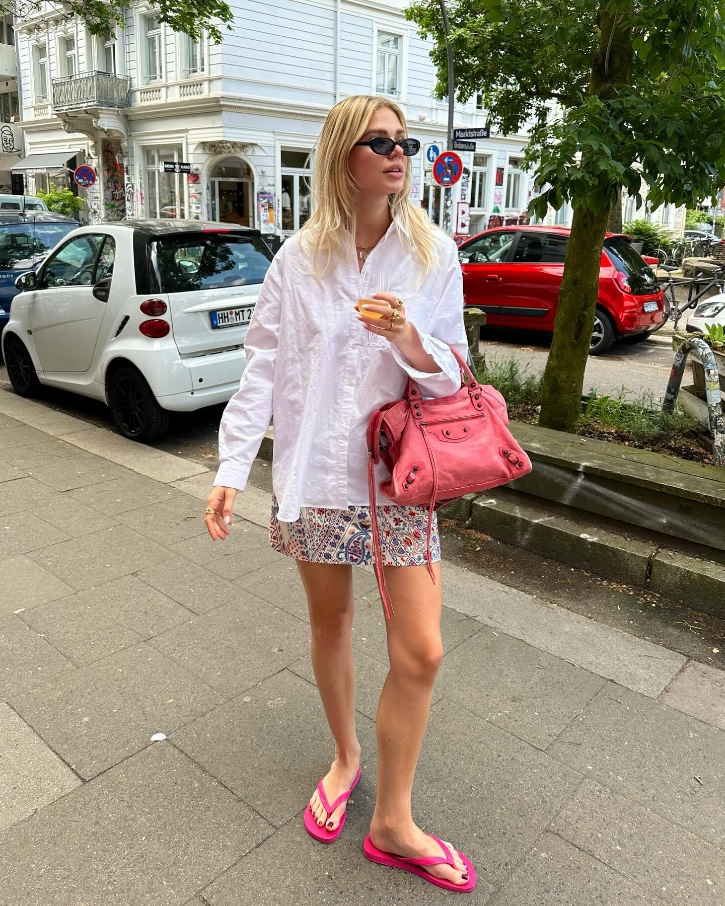 Modelo caminha com postura descontraída usando chinelo rosa vibrante, saia estampada, camisa branca ampla e bolsa rosa em