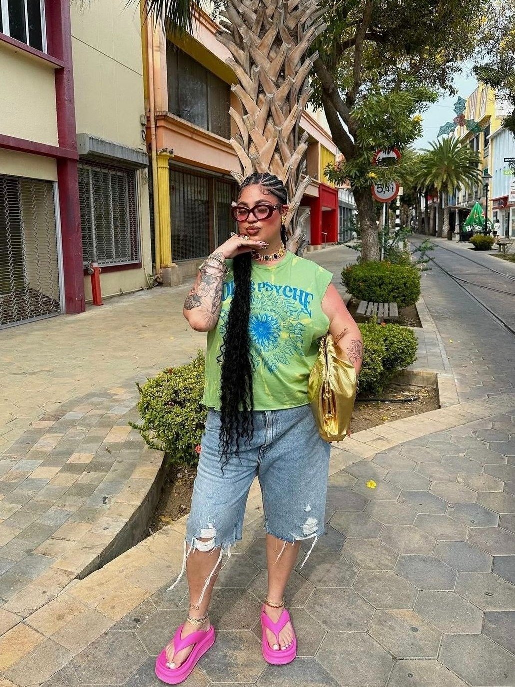 Mulher posa confiante com chinelo rosa de solado alto, bermuda jeans rasgada e camiseta verde limão estampada, em rua urbana.