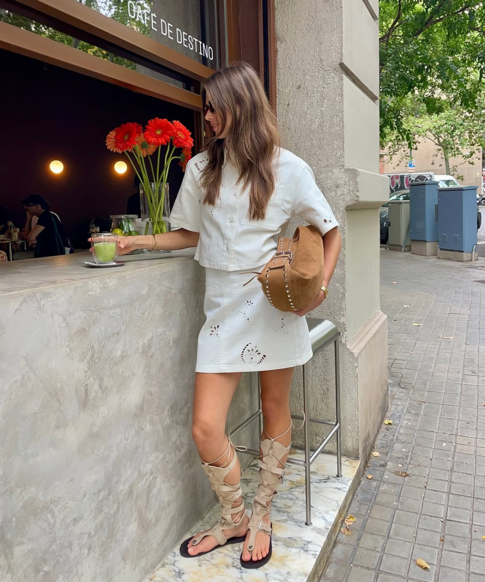 Modelo posa ao lado de balcão urbano usando sandália gladiadora bege com tiras largas, vestido branco com recortes e bolsa.