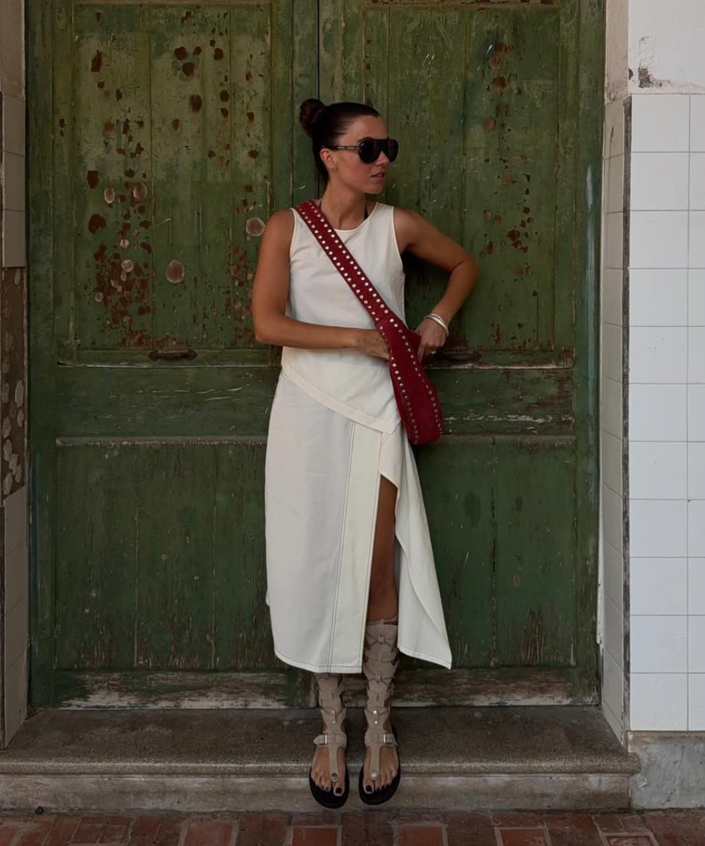 Modelo posa com vestido branco de modelagem assimétrica e sandália gladiadora bege em material sintético, combinando bolsa
