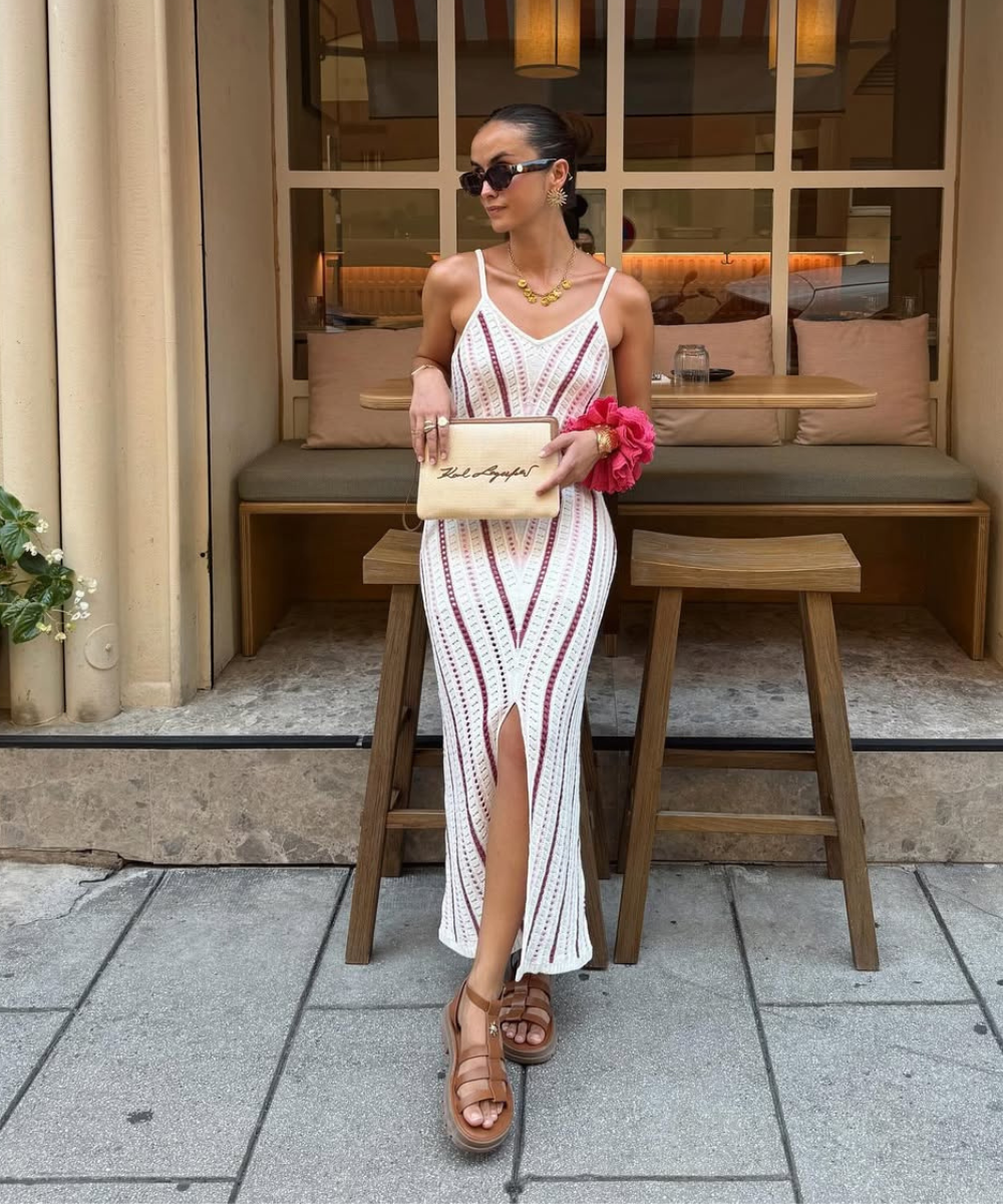 Modelo posa com vestido longo de tricô branco com listras vermelhas, clutch bege e sandália gladiadora marrom em couro.