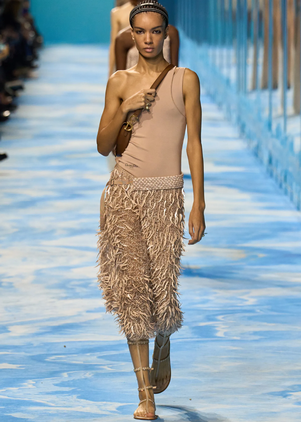 Modelo desfila com sandália gladiadora nude de tiras finas, combinada a top assimétrico e calça texturizada bege.