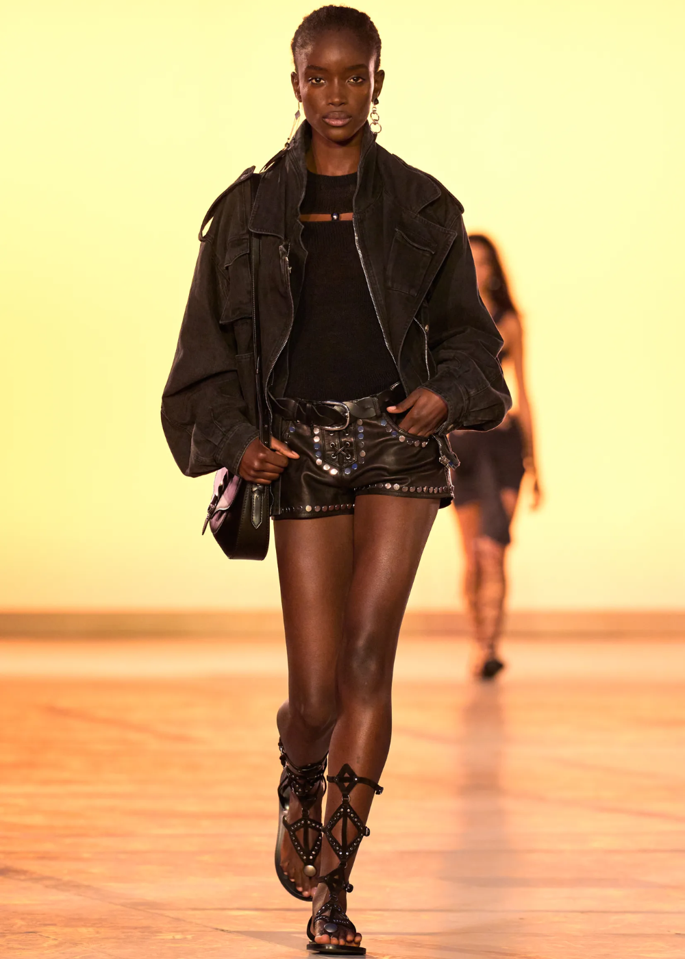 Modelo caminha com postura confiante usando sandália gladiadora preta com tiras geométricas, short e jaqueta oversized.