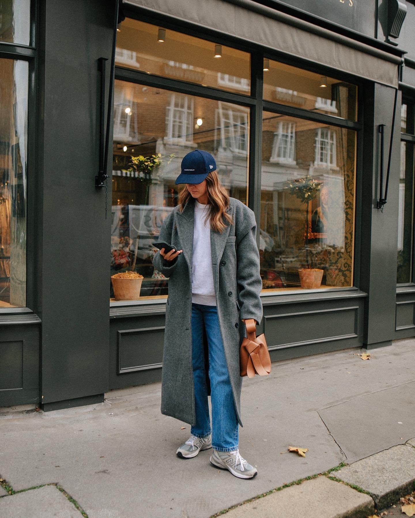 Modelo posa na calçada com sobretudo cinza oversized, jeans reto, tênis cinza esportivo e boné azul, segurando bolsa
