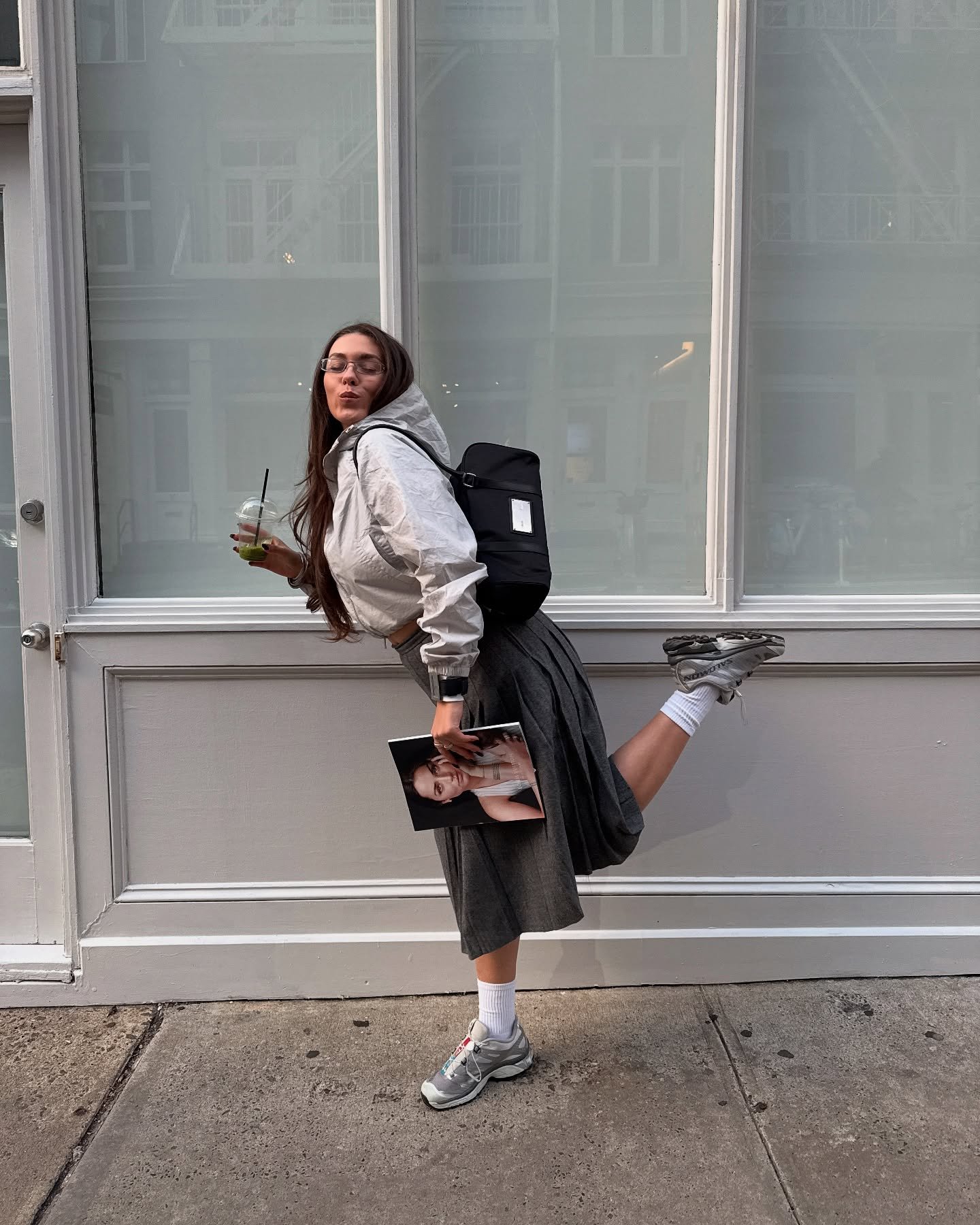 Mulher posa divertida com tênis cinza esportivo, saia midi plissada cinza, jaqueta cropped clara e mochila preta urbana.