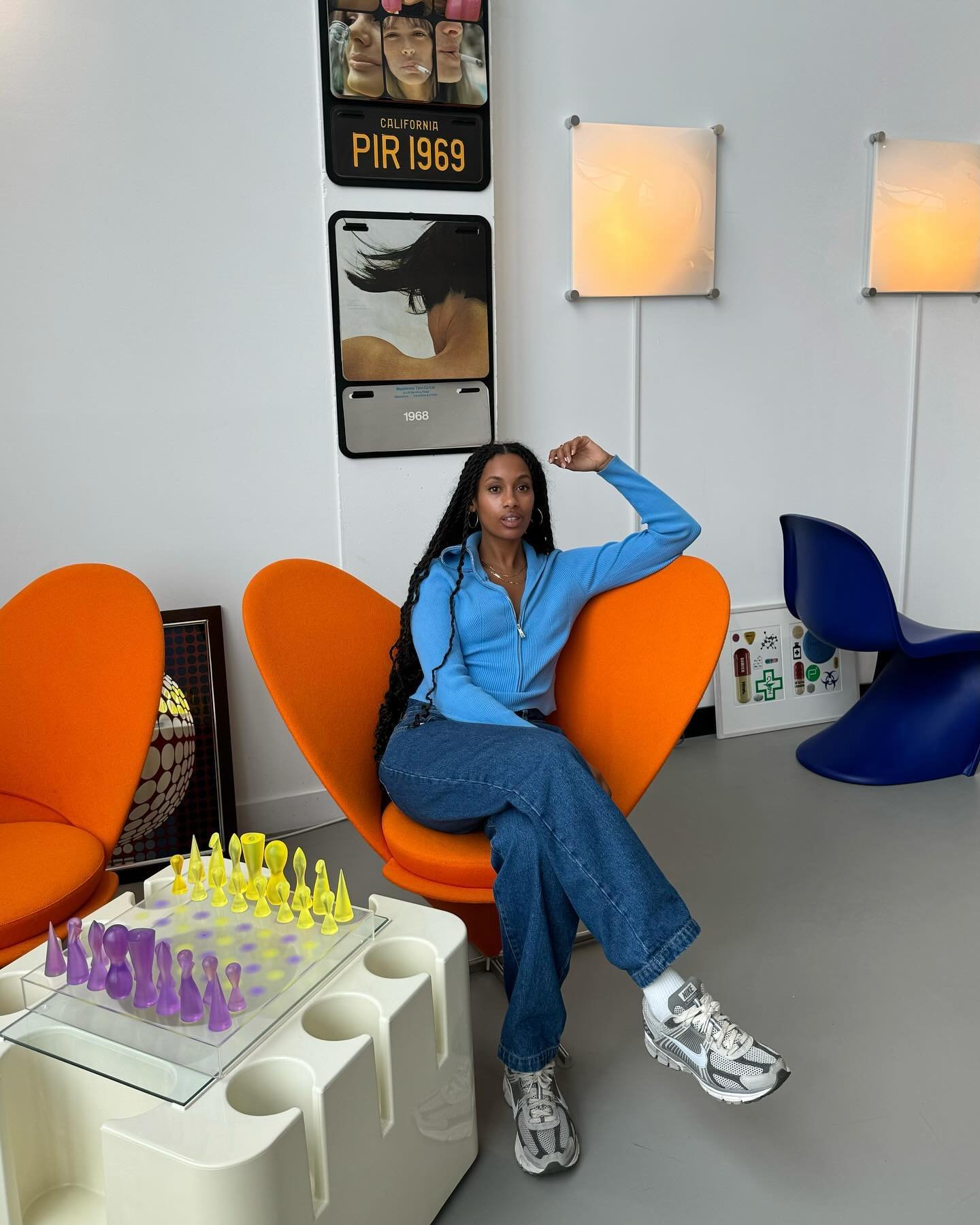 Modelo senta descontraída em cadeira laranja vibrante, veste blusa azul de tricô, jeans amplo e tênis cinza com detalhes em