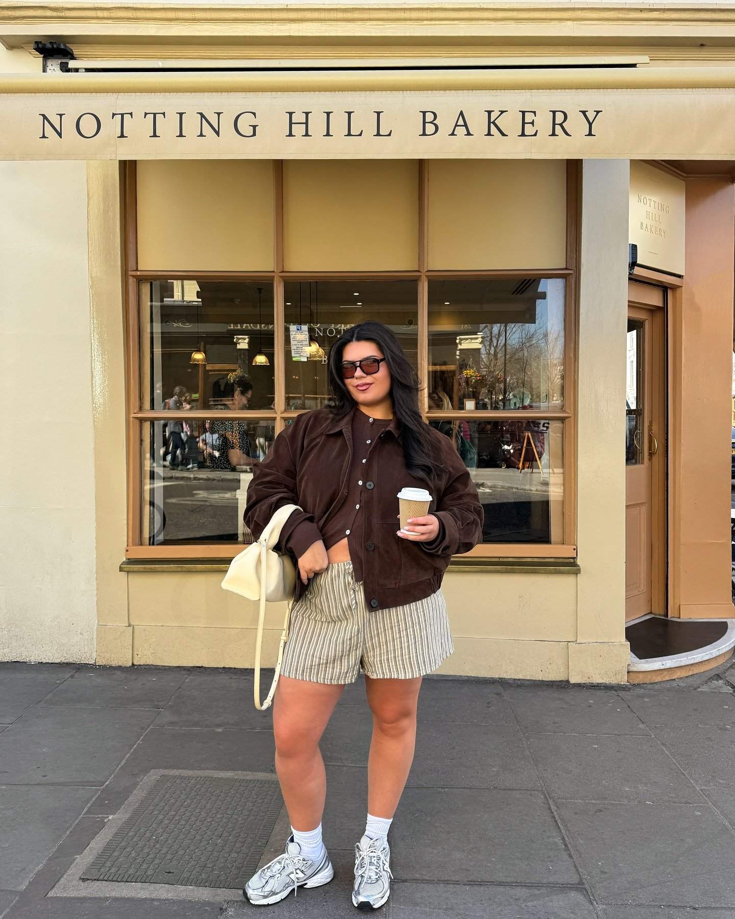 Modelo posa confiante em frente à padaria com tênis cinza esportivo, shorts listrados, jaqueta marrom e bolsa creme, clima
