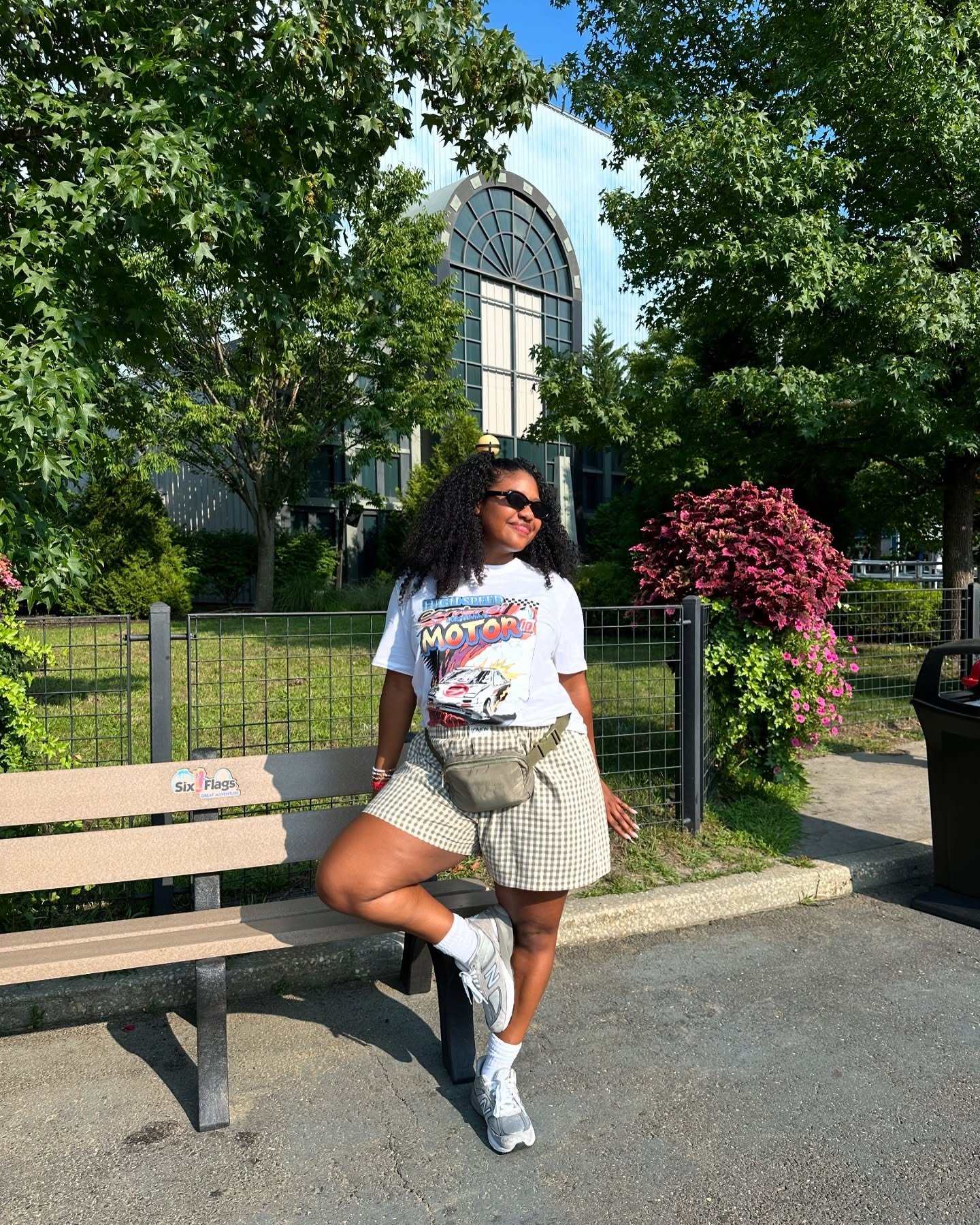 Mulher posa relaxada com looks confortáveis e leves em camiseta branca estampada, shorts xadrez e tênis cinza em parque