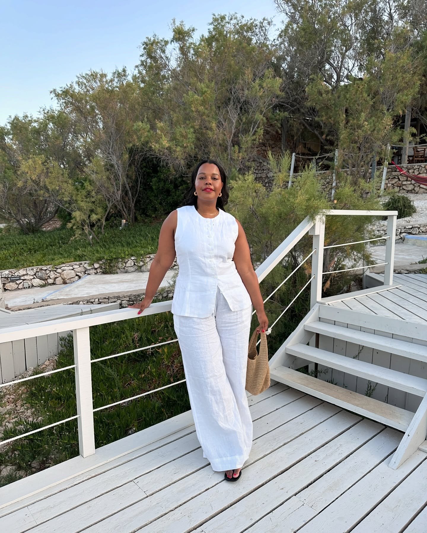 Mulher posa confiante com conjunto branco de linho fluido, regata e calça ampla, em cenário natural sereno, estilo cloud