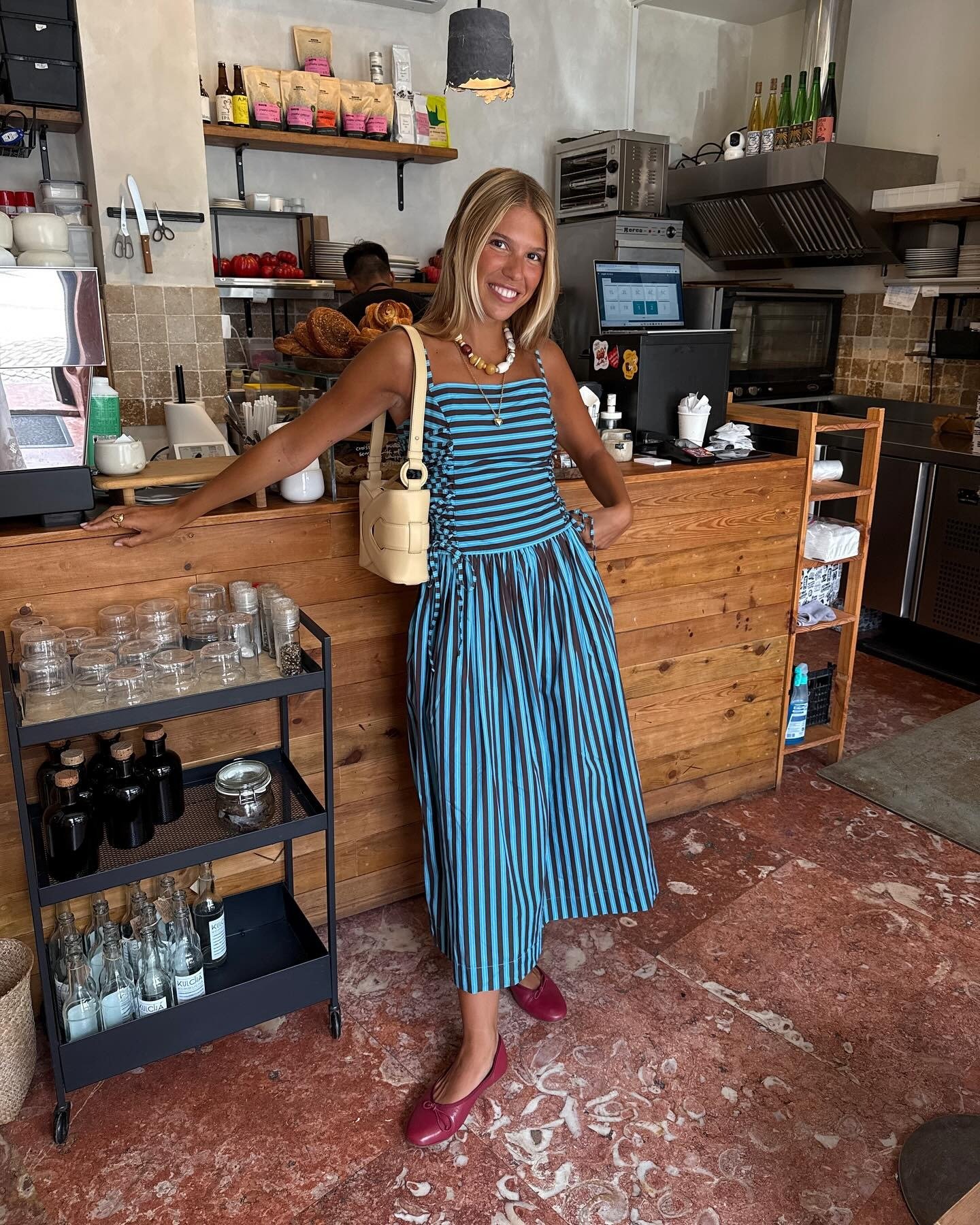 Modelo posa sorrindo com vestido midi listrado azul e preto, modelagem leve e fluida, bolsa bege e sapatilhas vinho, em café