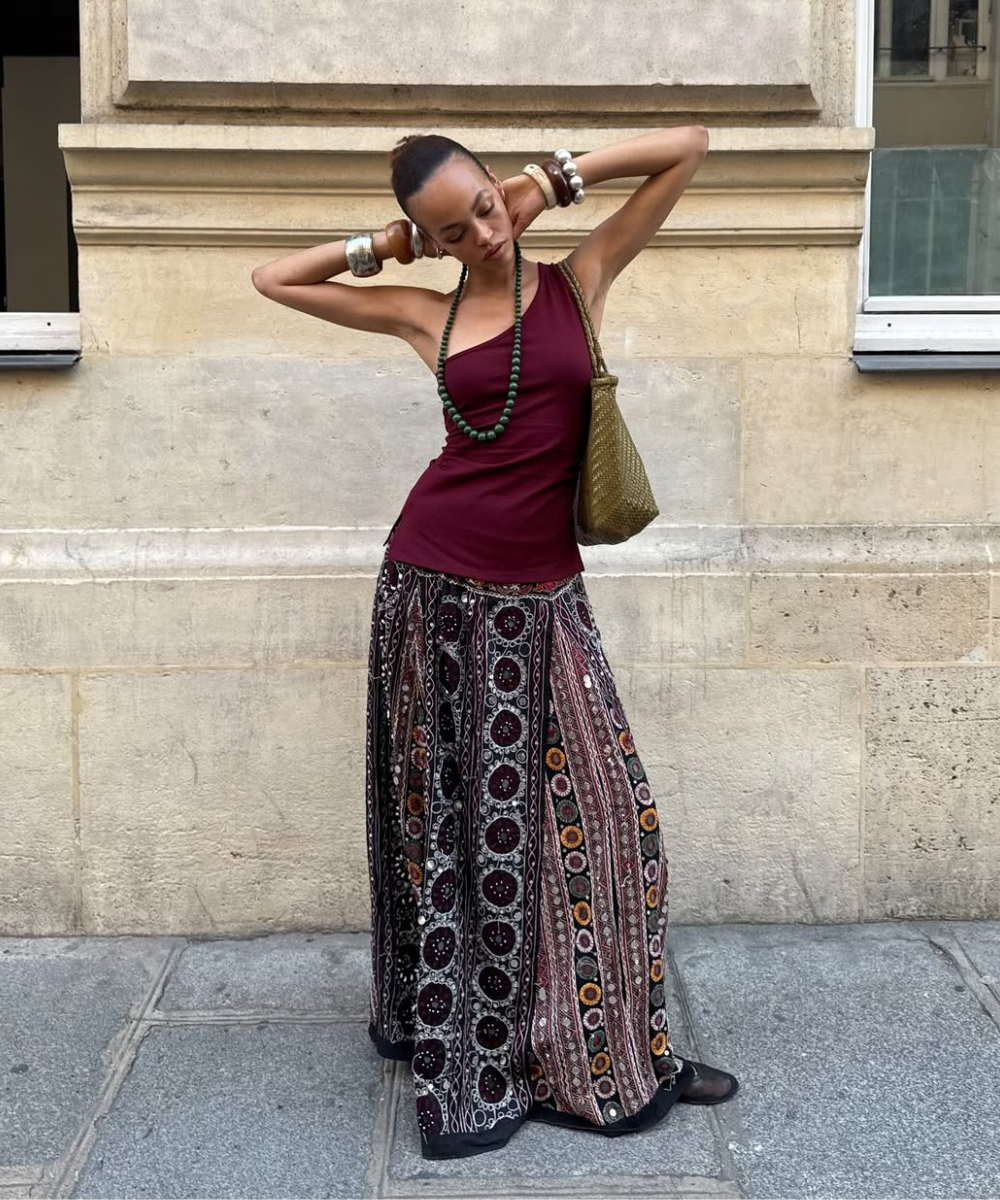 Modelo posa com regata vinho de alça única, saia longa estampada fluida, acessórios naturais e bolsa verde.
