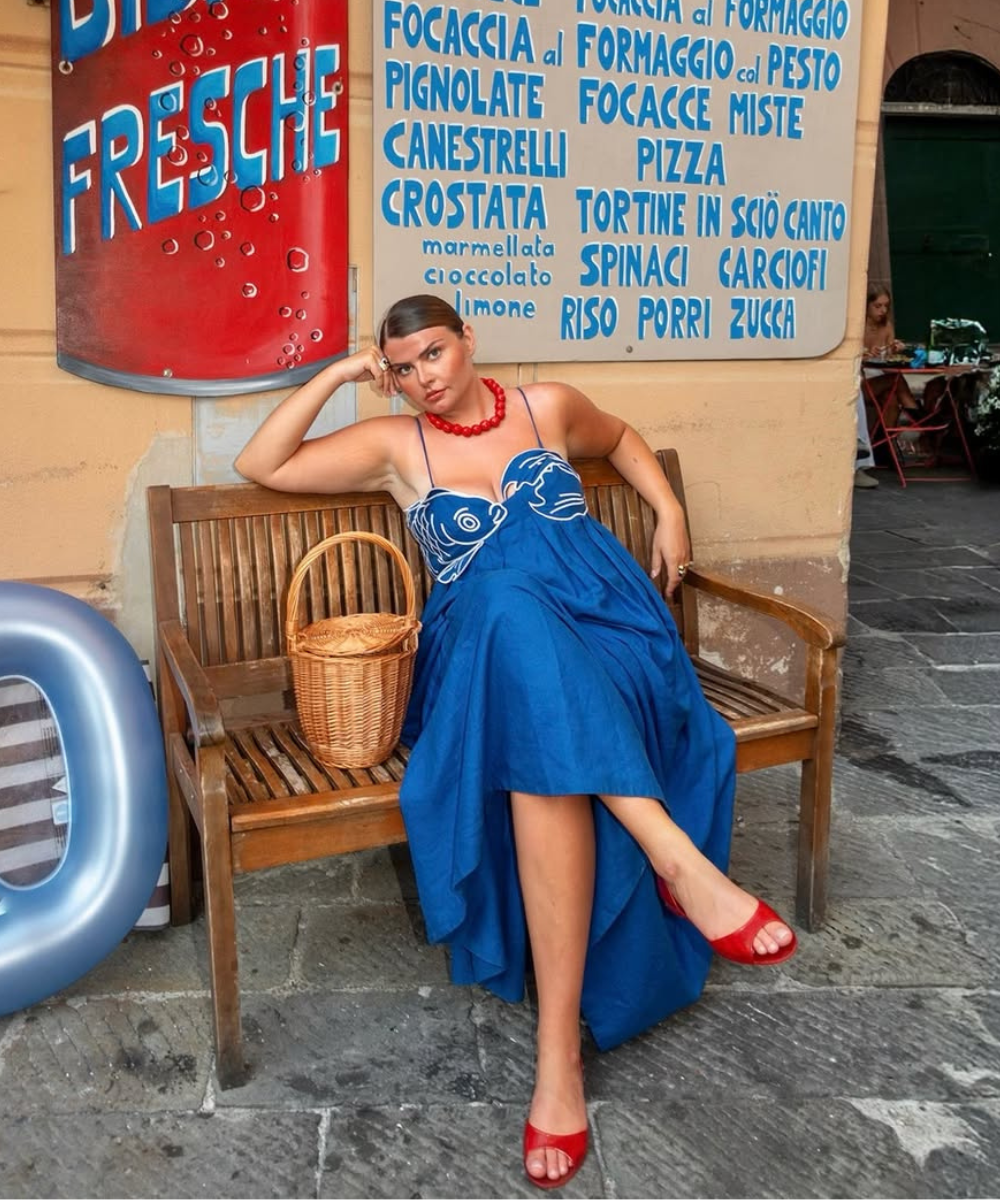 Modelo posa sentada com vestido azul de alças finas e estampa branca no busto, sandálias vermelhas, colar de contas e bolsa.