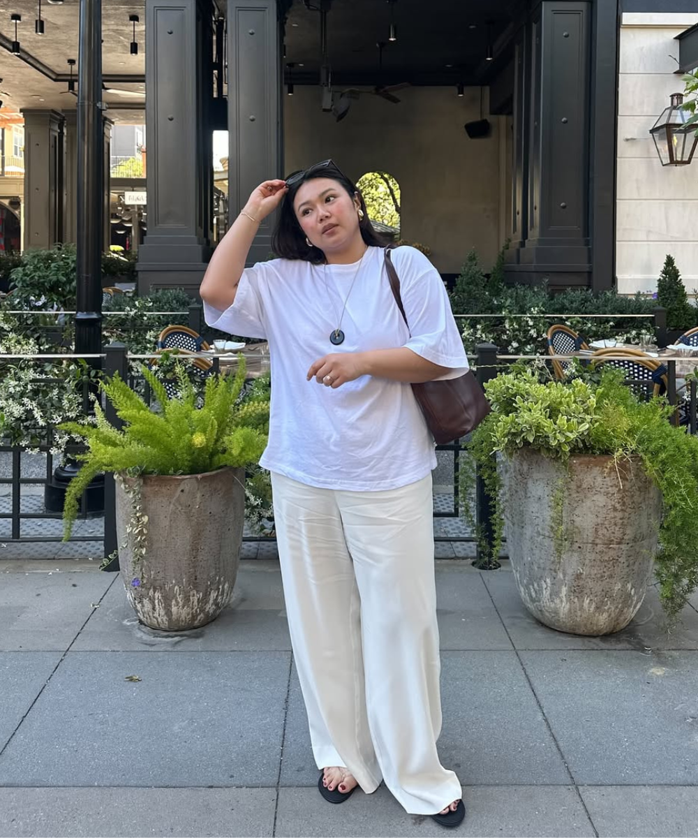 Modelo posa com looks de férias em tons neutros, usando camiseta branca ampla, calça pantalona fluida e sandálias pretas.