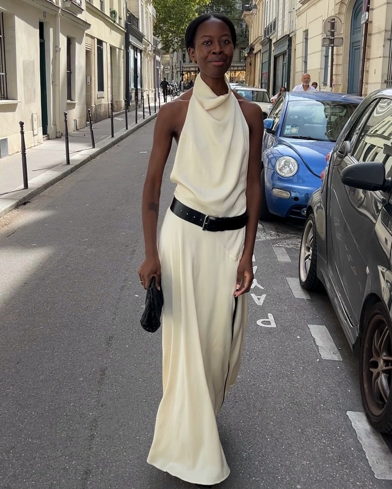 Modelo caminha confiante com vestido longo creme de tecido fluido, gola drapeada e cinto preto, em look elegante para o