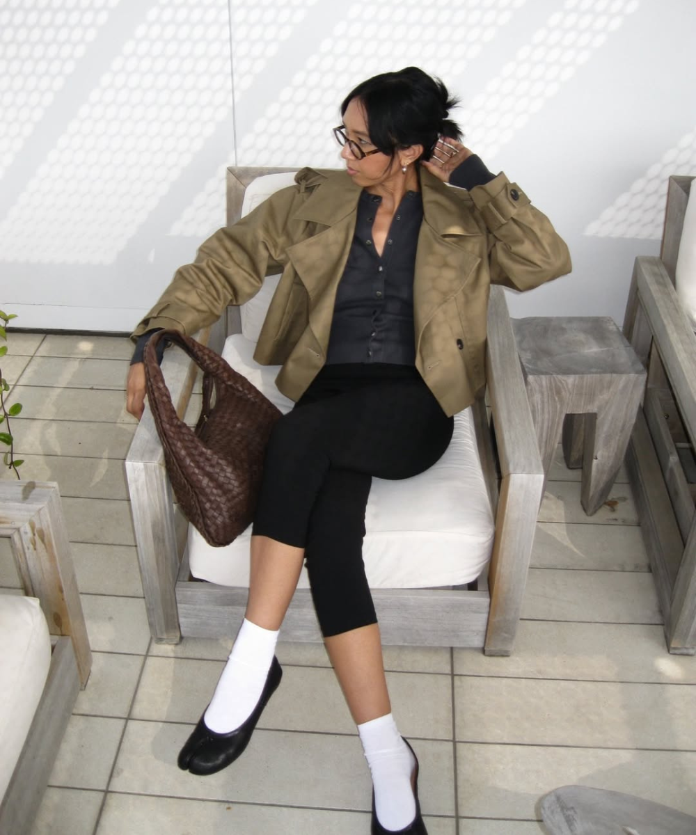 Mulher sentada com postura relaxada usa sapatilha com meia branca, calça preta cropped, camisa preta e jaqueta bege.