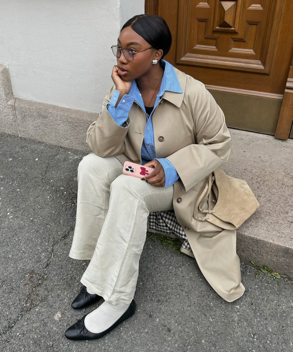 Mulher sentada com expressão pensativa usa sapatilha com meia branca, calça creme, camisa azul e sobretudo bege.