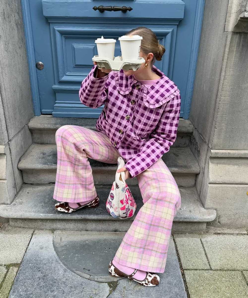Modelo senta em degrau urbano usando sapatilha com meia rosa, calça xadrez rosa e bege e jaqueta xadrez roxa com gola ampla.