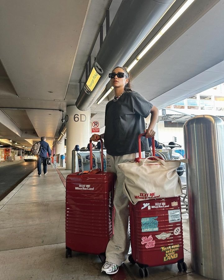 Modelo posa confiante em aeroporto com mala de viagem para as festas vermelha rígida, adesivos coloridos e bolsa creme