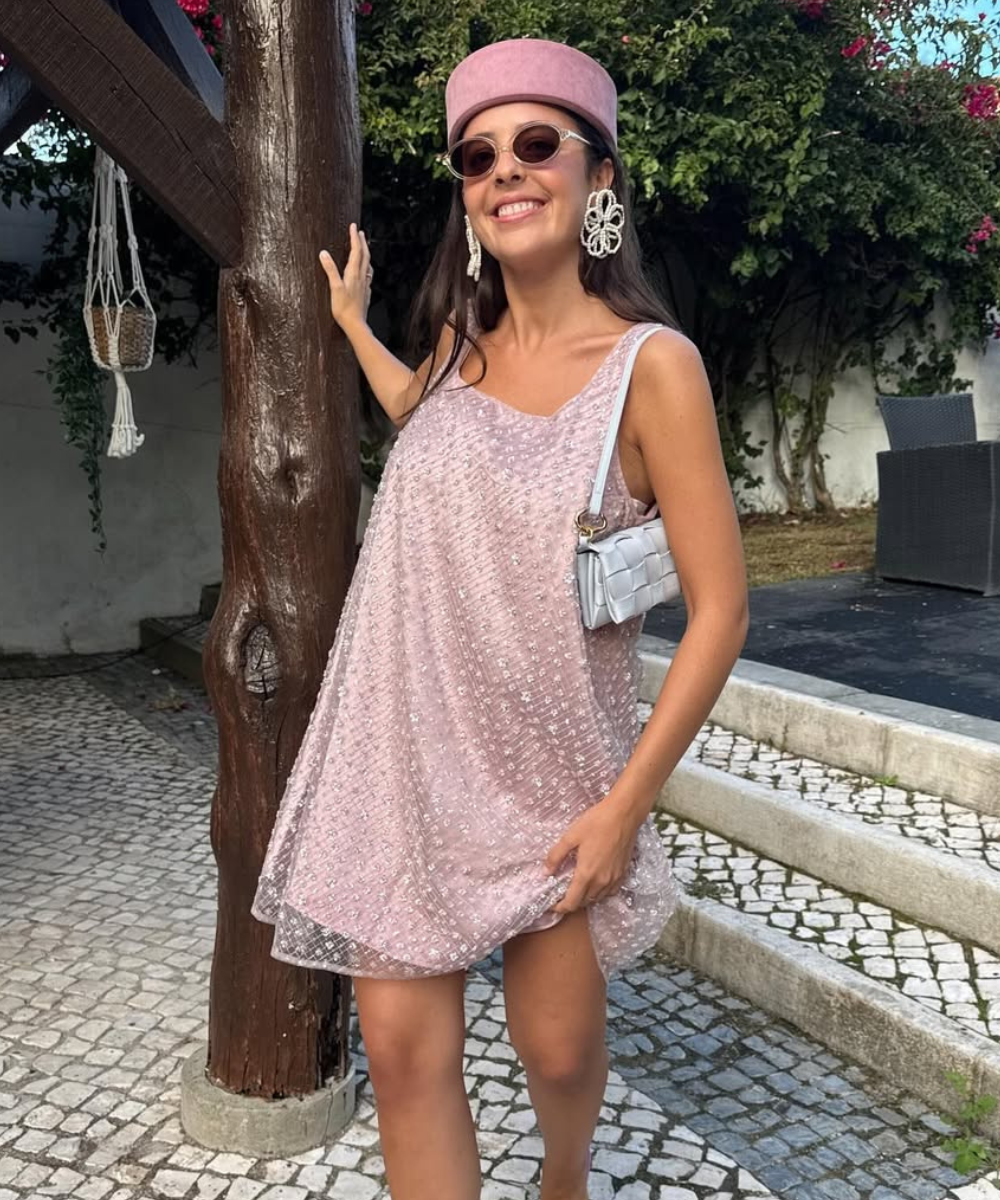 Modelo sorri e posa com vestido rosa claro texturizado, chapéu estruturado e bolsa branca entrelaçada.
