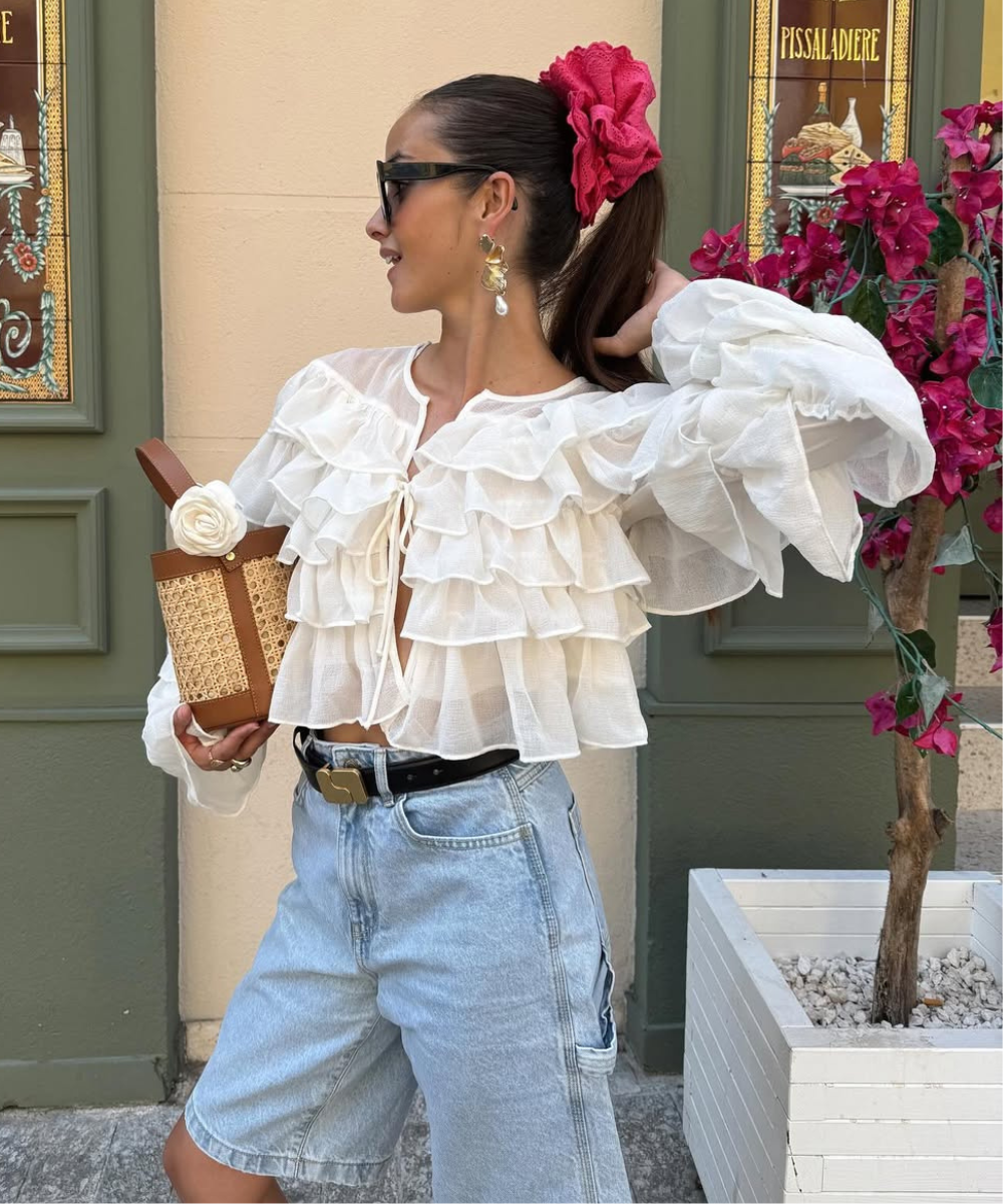 Modelo posa com blusa branca de babados em tecido leve, short jeans claro, bolsa de palha com alça marrom e flor branca.