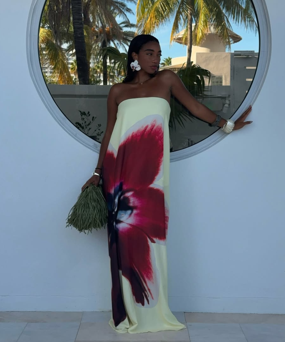 Modelo posa com vestido longo tomara que caia amarelo com estampa floral vermelha e acessório estiloso em formato de flor.