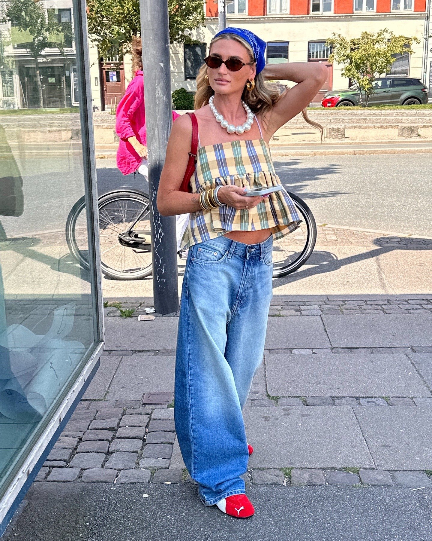 Modelo posa ao ar livre com top xadrez amarelo e azul de tecido leve, jeans largo, lenço azul, acessórios dourados e tênis